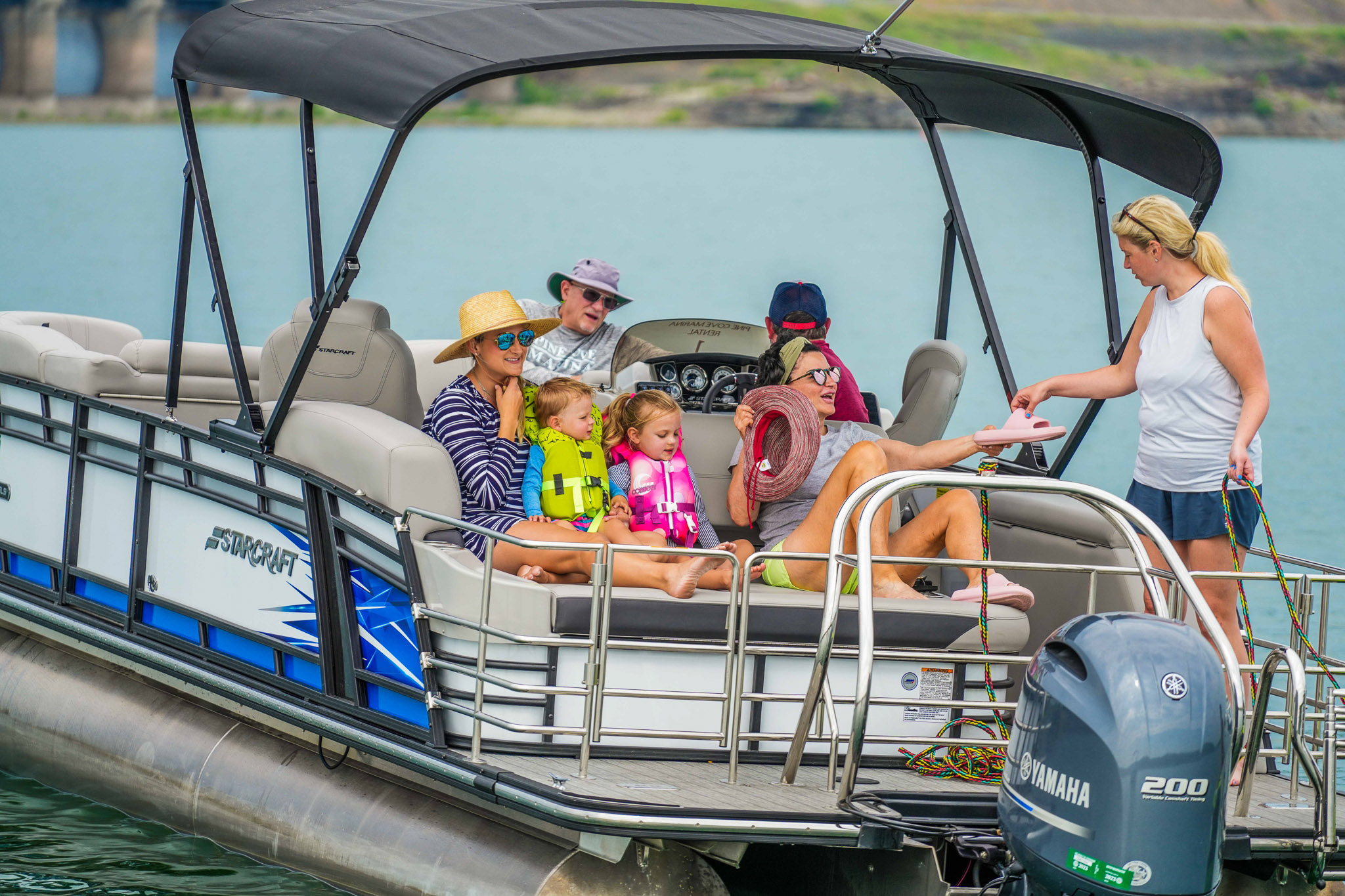 A group of people sits on a pontoon boat on an overcast Lake Tenkiller. Two children wear life jackets, and adults around them are in casual summer attire. Near the Bluewater Bay Marina dock, one person stands on the deck, engaging warmly with everyone aboard.