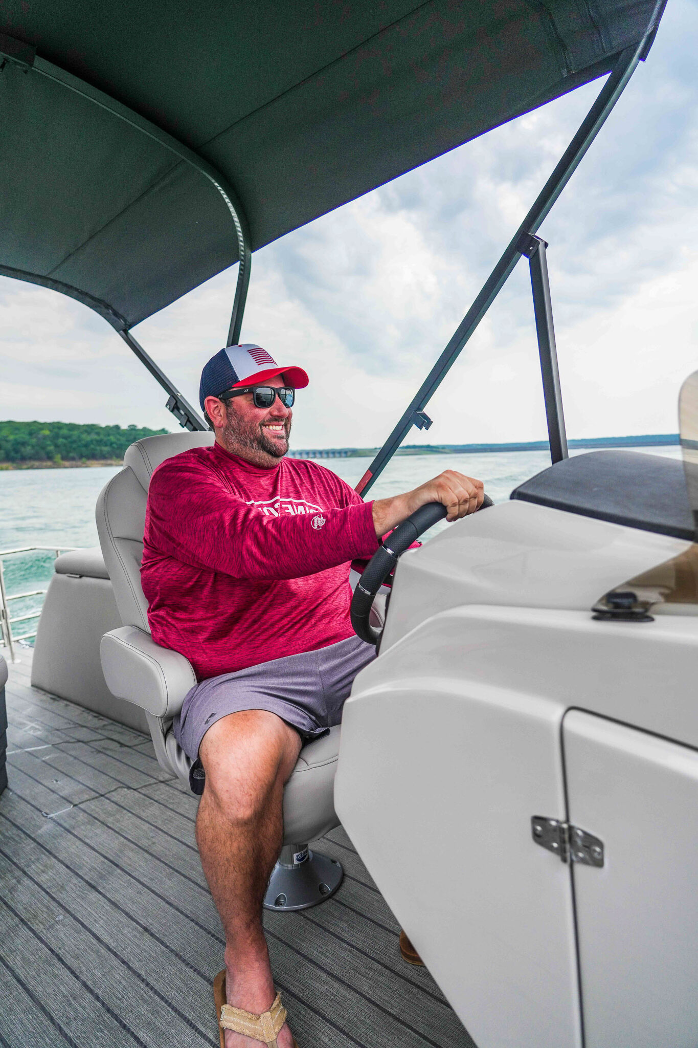 A man in a red long-sleeve shirt and cap happily drives a boat from Bluewater Bay Marina on Lake Tenkiller. Hes wearing sunglasses and shorts, with a canopy overhead. The water is calm, and trees are visible in the background under a cloudy sky.