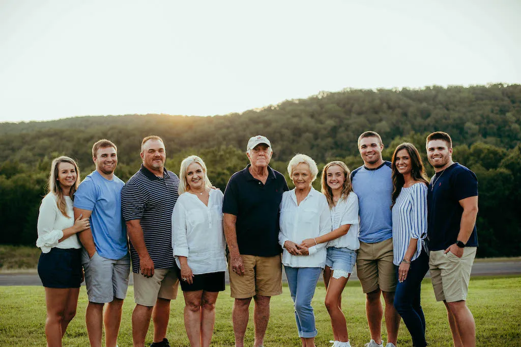 A group of people standing on grass with a forested hill in the background, at Bluewater Bay Marina. The group includes older and younger adults, all smiling and casually dressed. The sky is clear with the sun setting behind the hill near picturesque Lake Tenkiller.