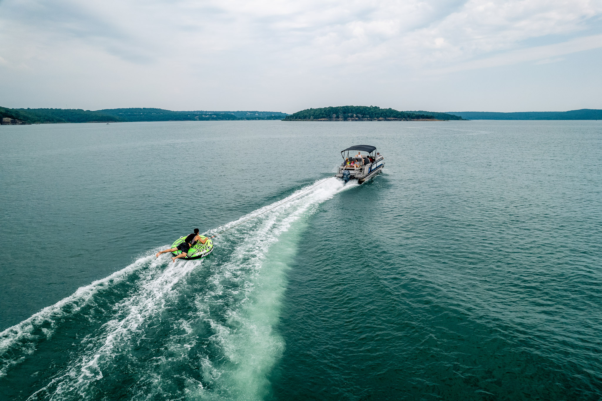 A person on a jet ski follows a motorboat across Lake Tenkillers vast, calm waters under a cloudy sky. The boat leaves a trail of white wake in the teal water, offering both excitement and tranquility. Distant hills are visible on the horizon, beckoning adventurers from Bluewater Bay Marina.