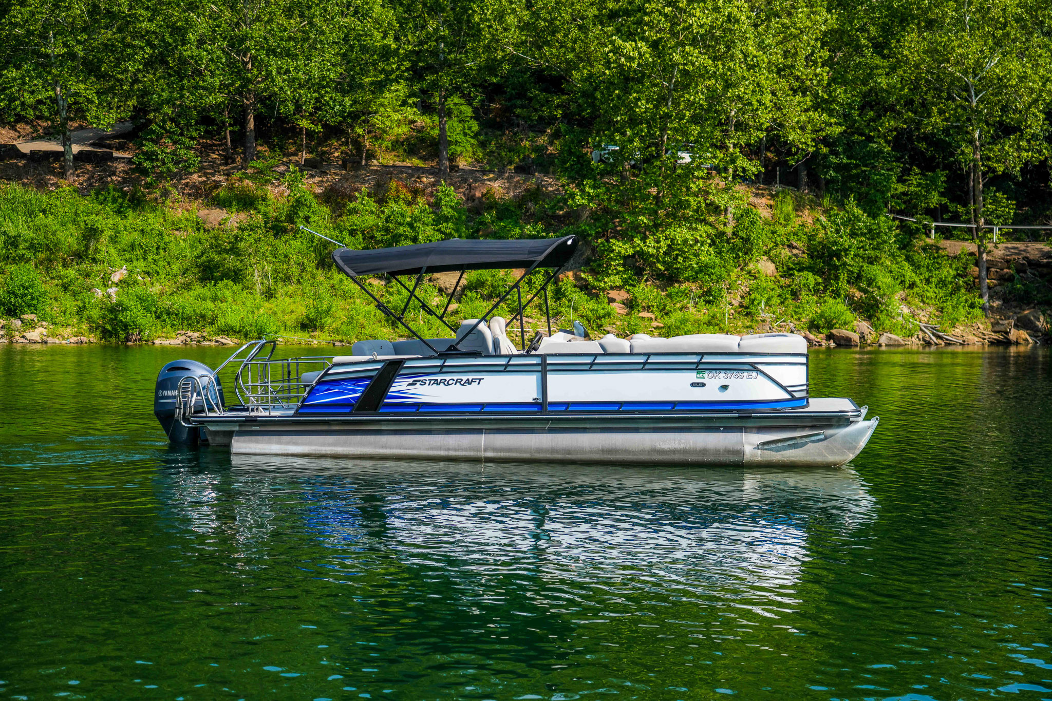A blue and white pontoon boat with a black canopy floats on the serene waters of Lake Tenkiller, surrounded by lush green trees lining the shore, just a short distance from Bluewater Bay Marina.