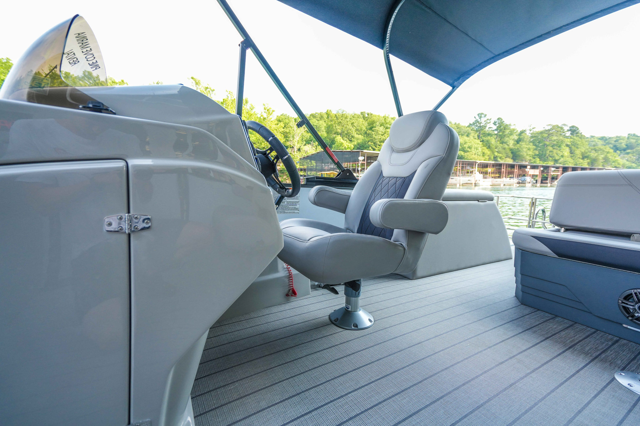 The captains seat on the boat deck under a blue canopy, with a steering wheel and controls to the left, offers views of Lake Tenkiller. The gray deck overlooks serene waters framed by green trees and dock structures, perfect for your next boat rental adventure.