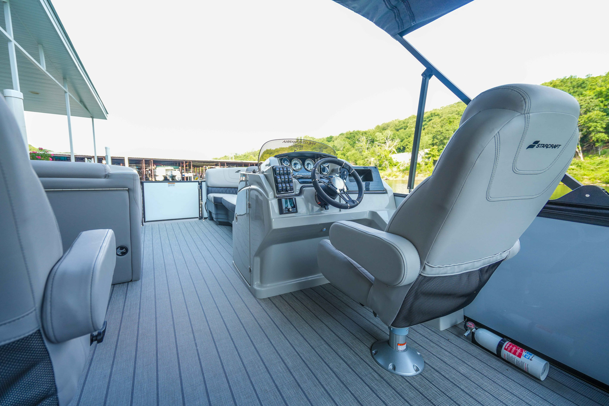 Interior of a pontoon boat featuring the captains chair and control panel, perfect for exploring Lake Tenkiller. The boat floor is covered with gray planks, while a fire extinguisher is mounted below. In the background, lush greenery and part of Bluewater Bay Marinas dock structure are visible.