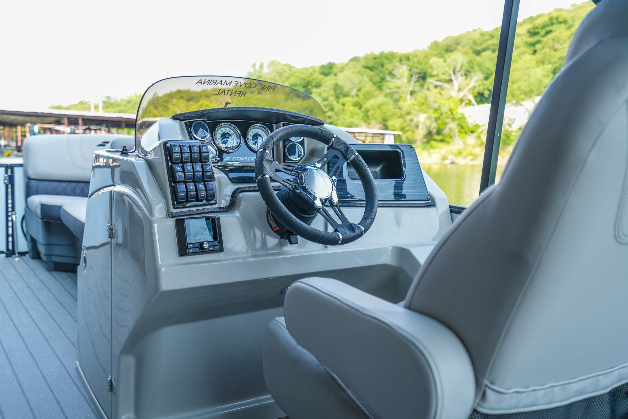 A modern boat steering console with a wheel, control panel, and gauges is shown. The console is light gray with a cushioned captains seat. A body of water and lush green trees are visible in the background—perfect for exploring Lake Tenkiller or setting off from Bluewater Bay Marina.