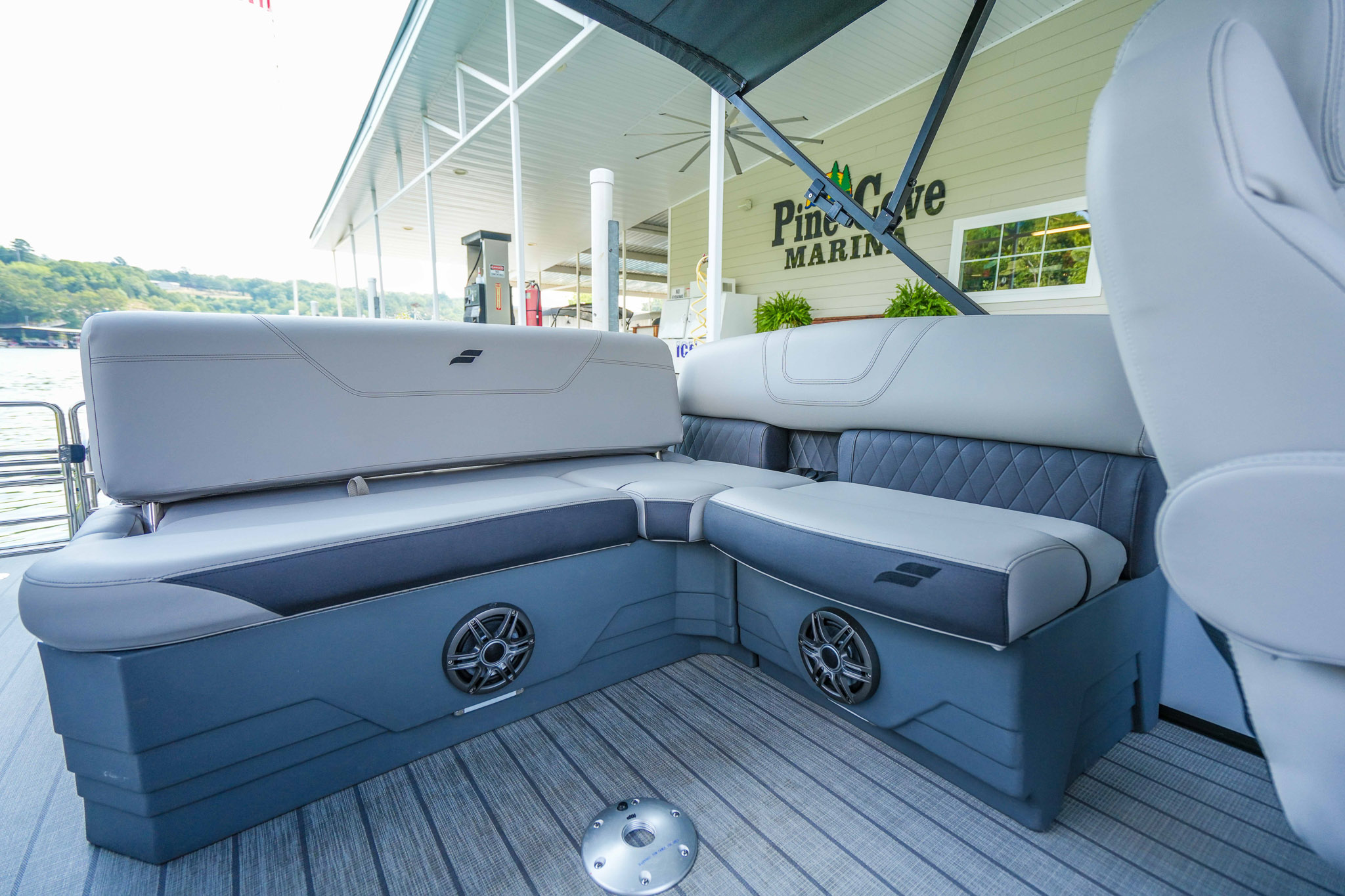 Luxury boat interior with gray cushioned seating and sleek design. Speakers are installed under the seats. The backdrop shows a marina with signage that reads Pine Cove Marina at Lake Tenkiller, surrounded by lush greenery, offering an idyllic boat rental experience.