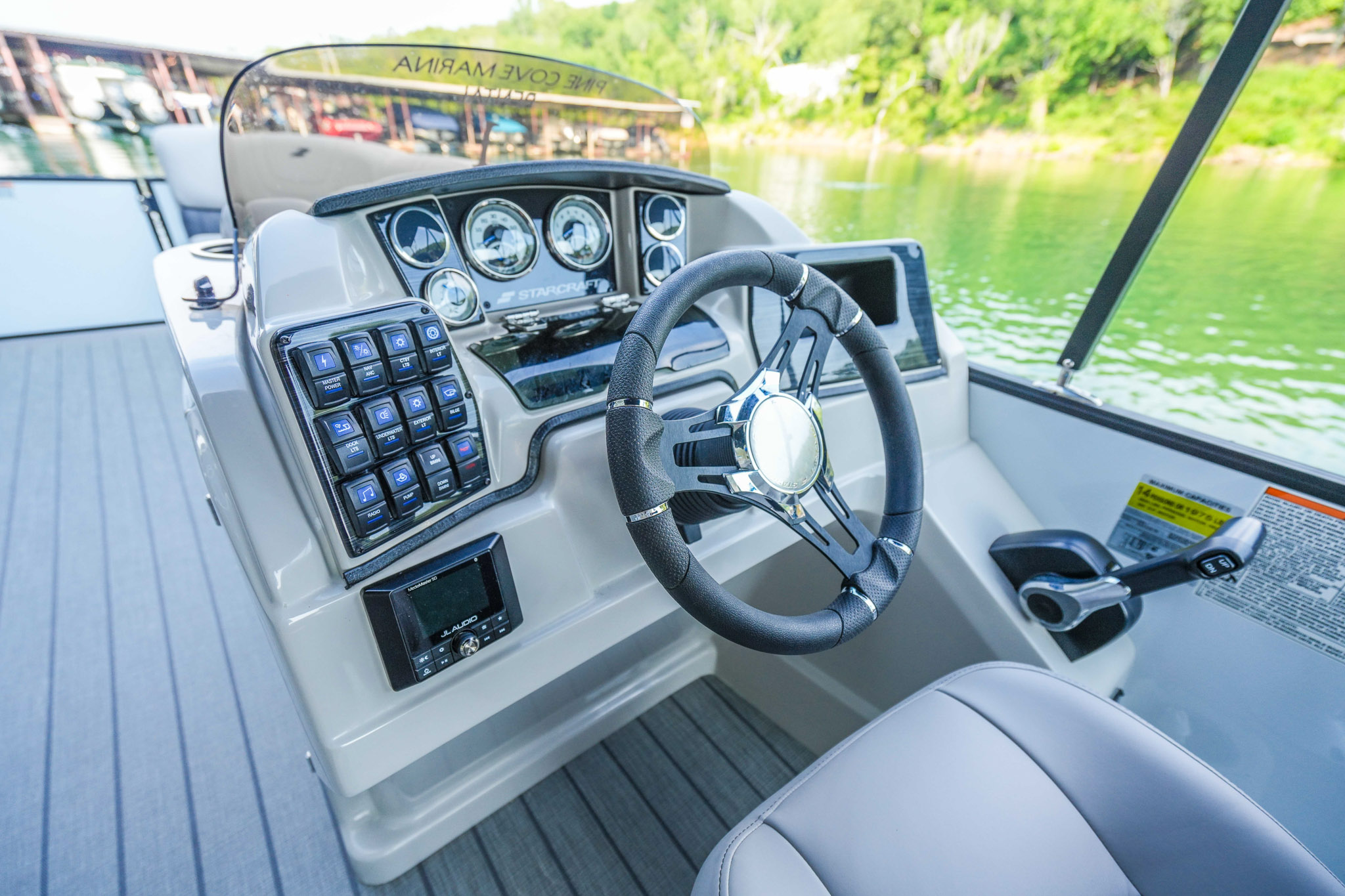 Close-up of a boats steering wheel and control panel with various gauges and switches, set against the serene backdrop of Bluewater Bay Marina. The surrounding calm river and lush greenery complement the wooden-textured floor.
