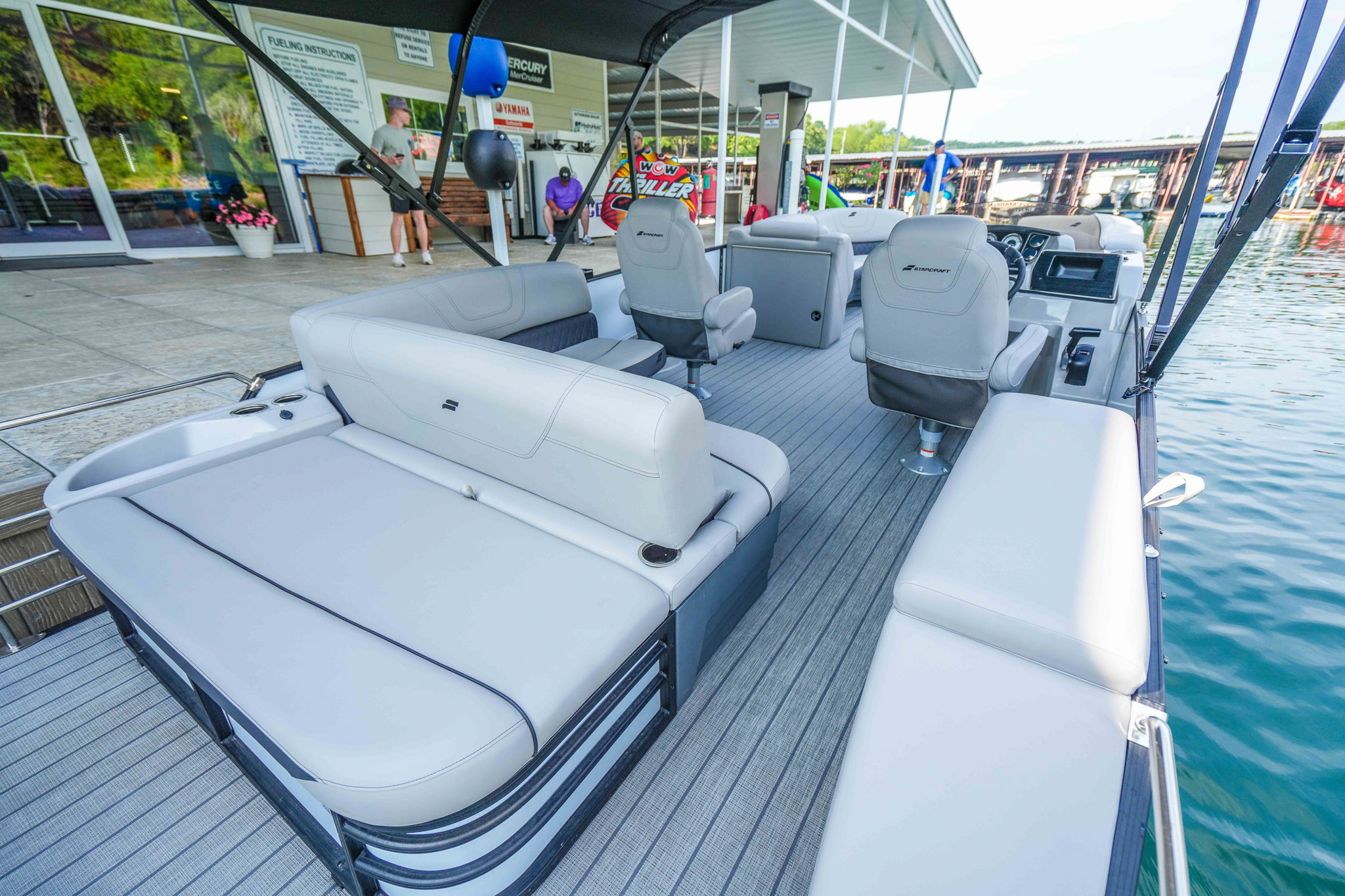 The interior of a luxury pontoon boat, featuring light gray seating and a modern design, is docked at Bluewater Bay Marina under a clear sky. The calm waters of Lake Tenkiller glisten to the right, offering an ideal setting for an unforgettable boat rental experience.