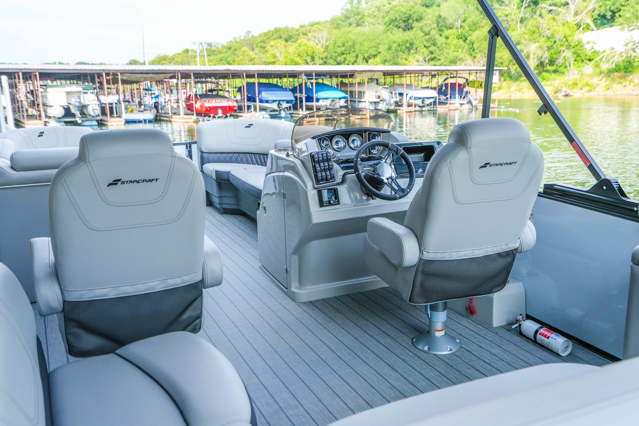 The image showcases the interior of a boat from Bluewater Bay Marina, featuring two white captains chairs at the helm with a control panel and steering wheel. The deck is surrounded by cushioned seating, and a lakeside dock at Lake Tenkiller with boats and trees is visible in the background.