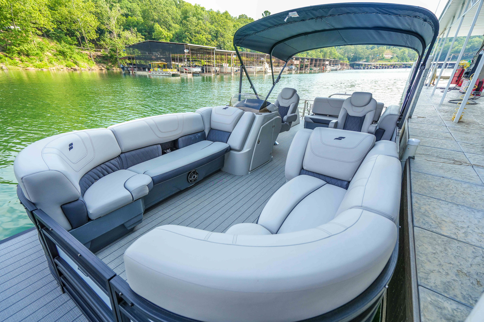 Pontoon boat with gray seating and a covered canopy, docked on the serene Lake Tenkiller. Enveloped by lush green trees and a dock in the background, this idyllic spot is perfect for your next adventure with a convenient boat rental from Bluewater Bay Marina.