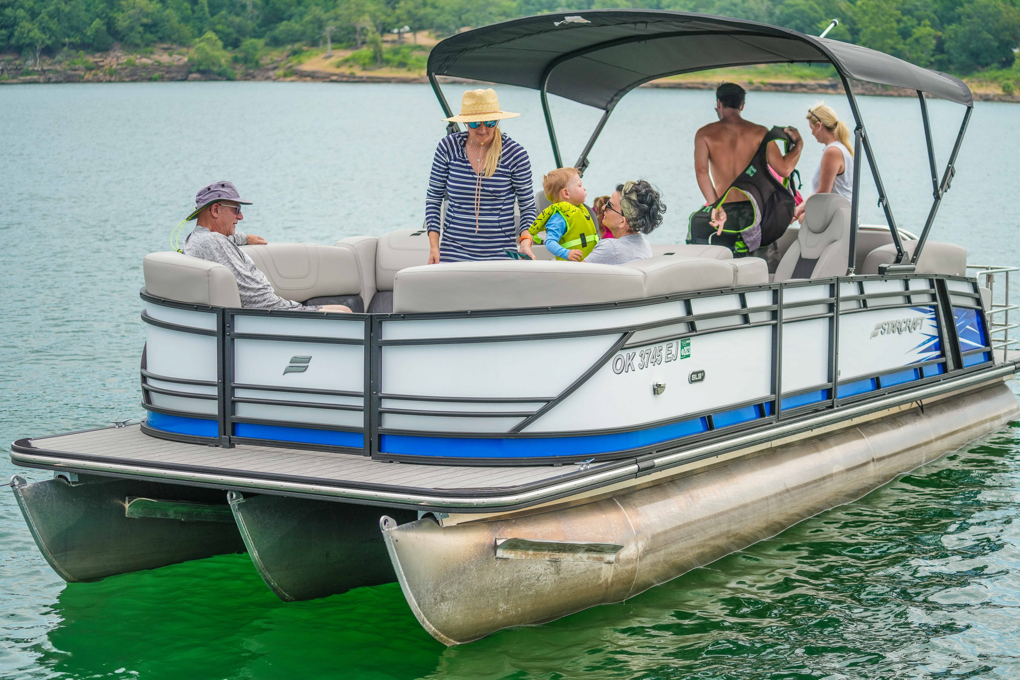 A group of people, including adults and children, enjoy a sunny day on a pontoon boat from Bluewater Bay Marina. The boat glides across Lake Tenkillers calm waters, with trees lining the shore. Some passengers sit while others stand and interact on this delightful boat rental adventure.