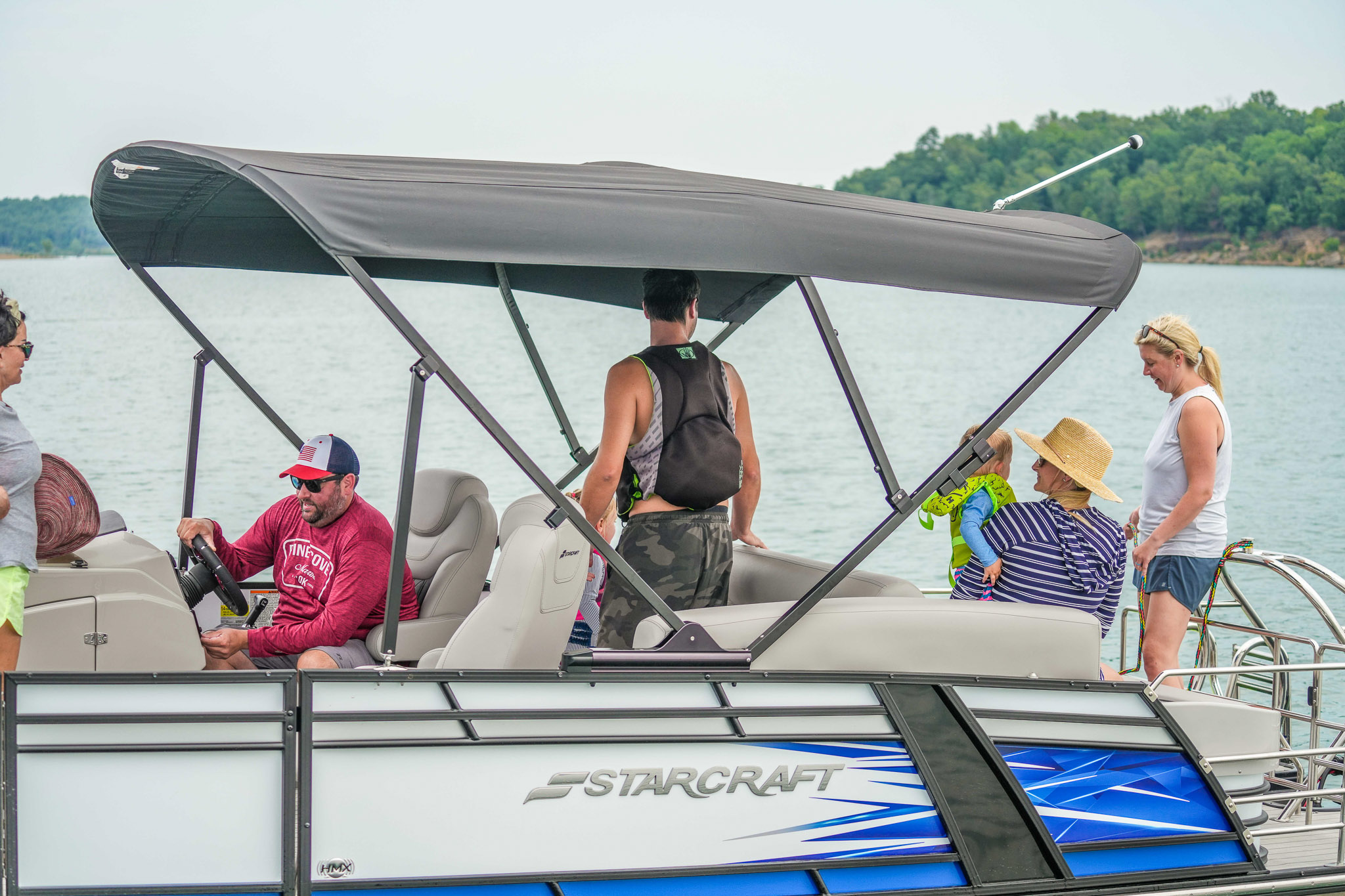 A group of people relax on a pontoon boat with a canopy at Lake Tenkiller. One person steers, while others chat and enjoy the outing. Trees line the shore in the background under a cloudy sky, near Bluewater Bay Marinas popular boat rental spot.
