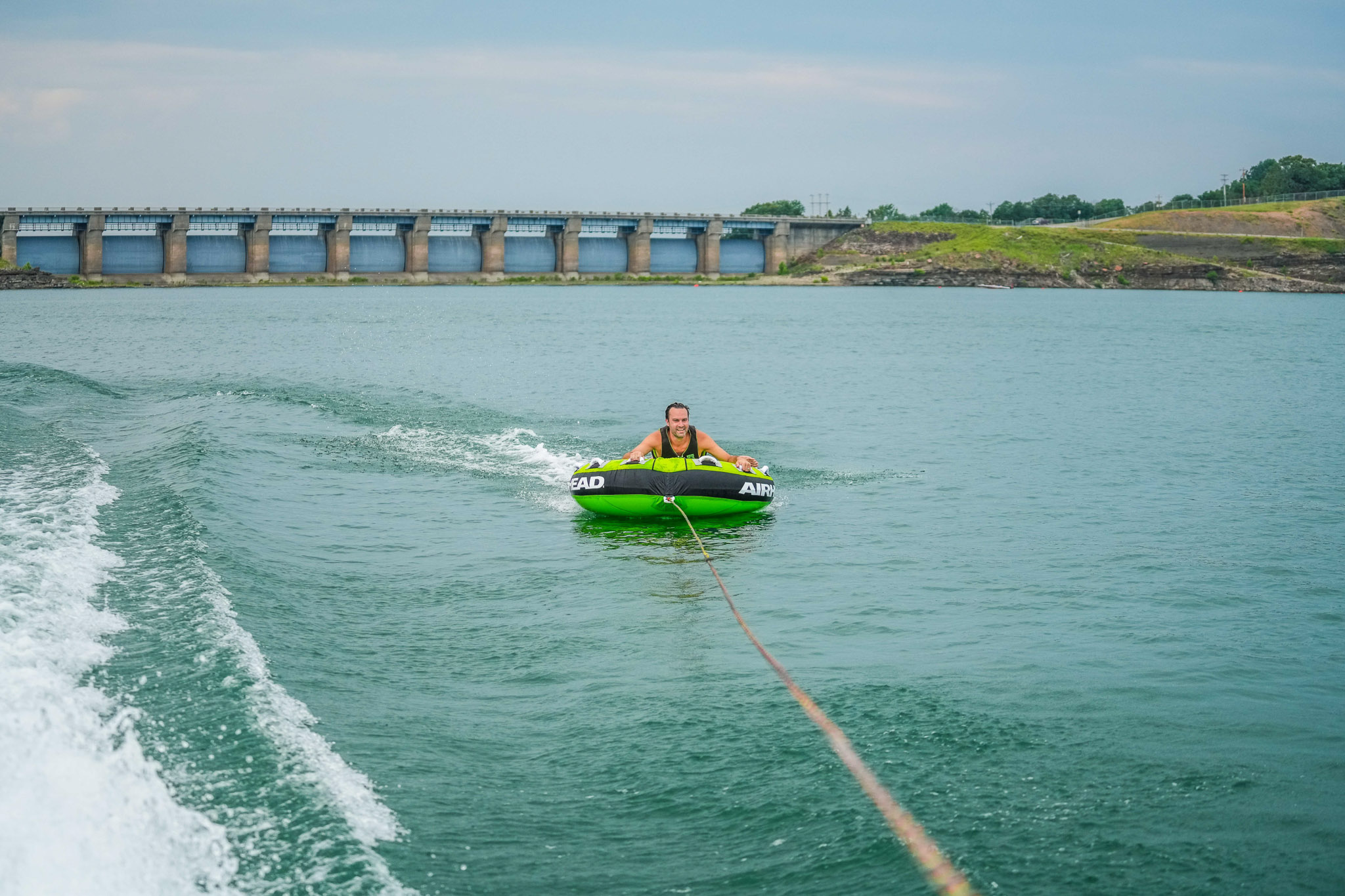 A person is enjoying a thrilling ride on a green inflatable tube towed by a boat on Lake Tenkiller. They glide past the dam and lush greenery of Bluewater Bay Marina, all under the watchful eye of a cloudy sky.