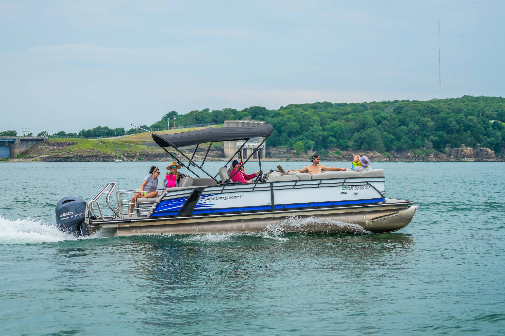 A group of people are enjoying a sunny day on a pontoon boat rented from Bluewater Bay Marina, cruising on Lake Tenkiller. The boat has a canopy, and the calm water reflects the blue sky. In the background, lush green trees frame the distant shoreline.
