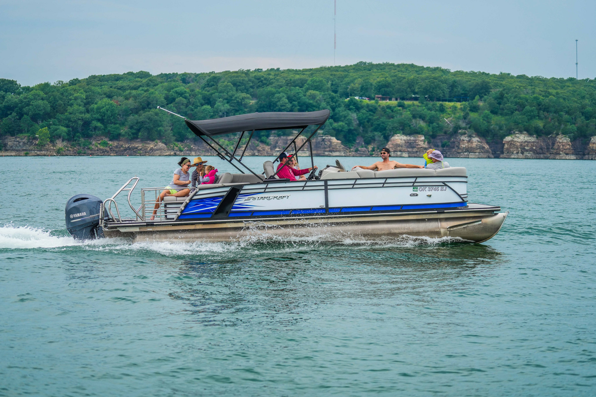 A pontoon boat from Bluewater Bay Marina with three people onboard cruises quickly on Lake Tenkiller, creating waves beneath its canopy. Forested hills stand proudly in the background under a clear sky.