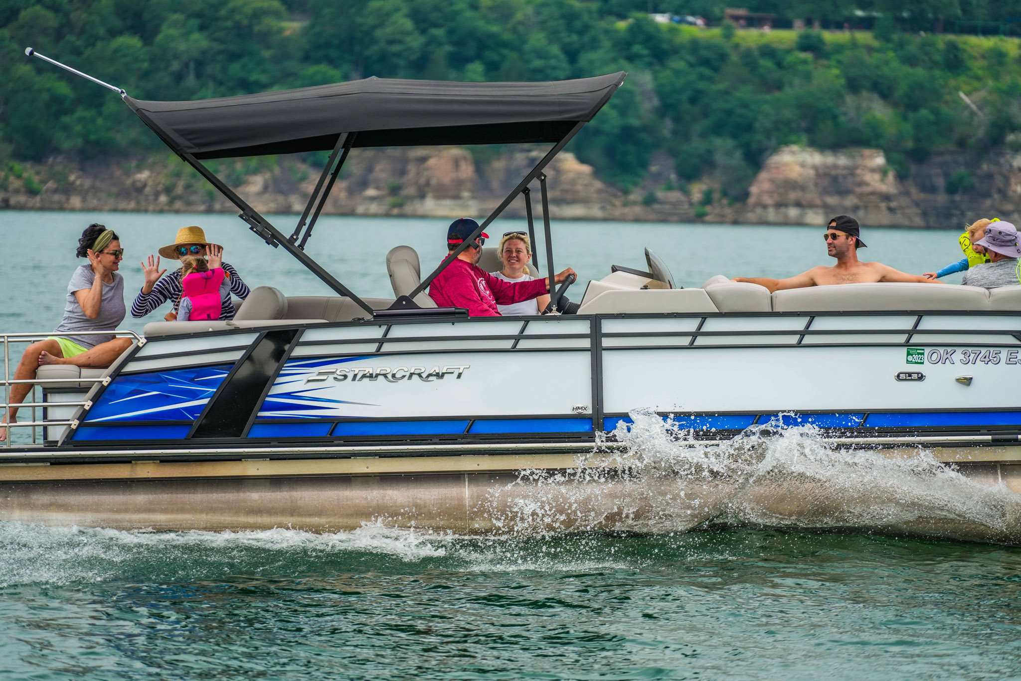 A group of people enjoys a boat ride on a pontoon in scenic Lake Tenkiller. Some chat while one steers as waves splash against the boat. Lush green trees and rocky terrain form the backdrop, creating the perfect day courtesy of Bluewater Bay Marinas boat rental service.
