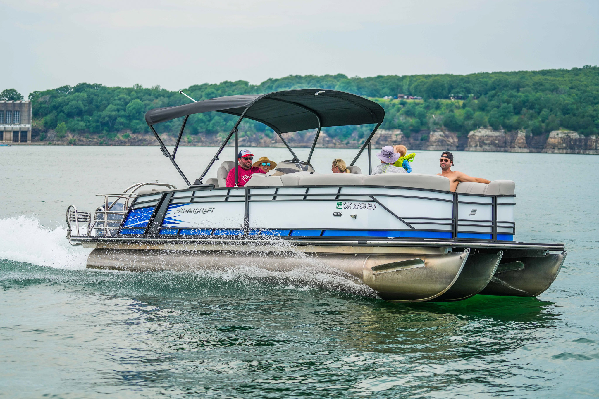 A group of people enjoying a ride on a pontoon boat with a black canopy, rented from Bluewater Bay Marina, moves swiftly through Lake Tenkiller. Water splashes against the hull as trees and rocky cliffs frame the cloudy sky in the background.