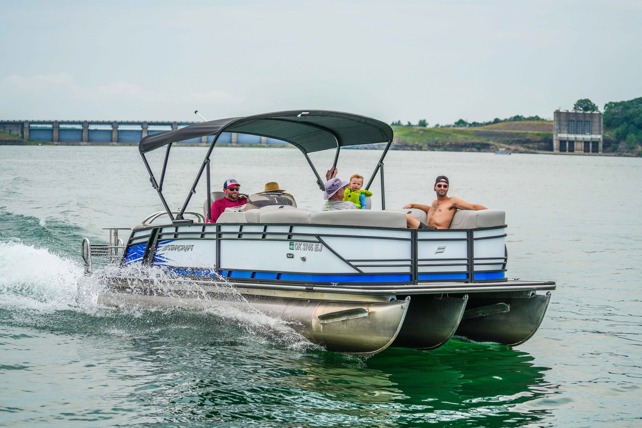 A group of people enjoy a ride on a pontoon boat rented from Bluewater Bay Marina in Lake Tenkiller. One person steers while others relax on the boats seats. A bridge and trees are visible in the background under a cloudy sky.