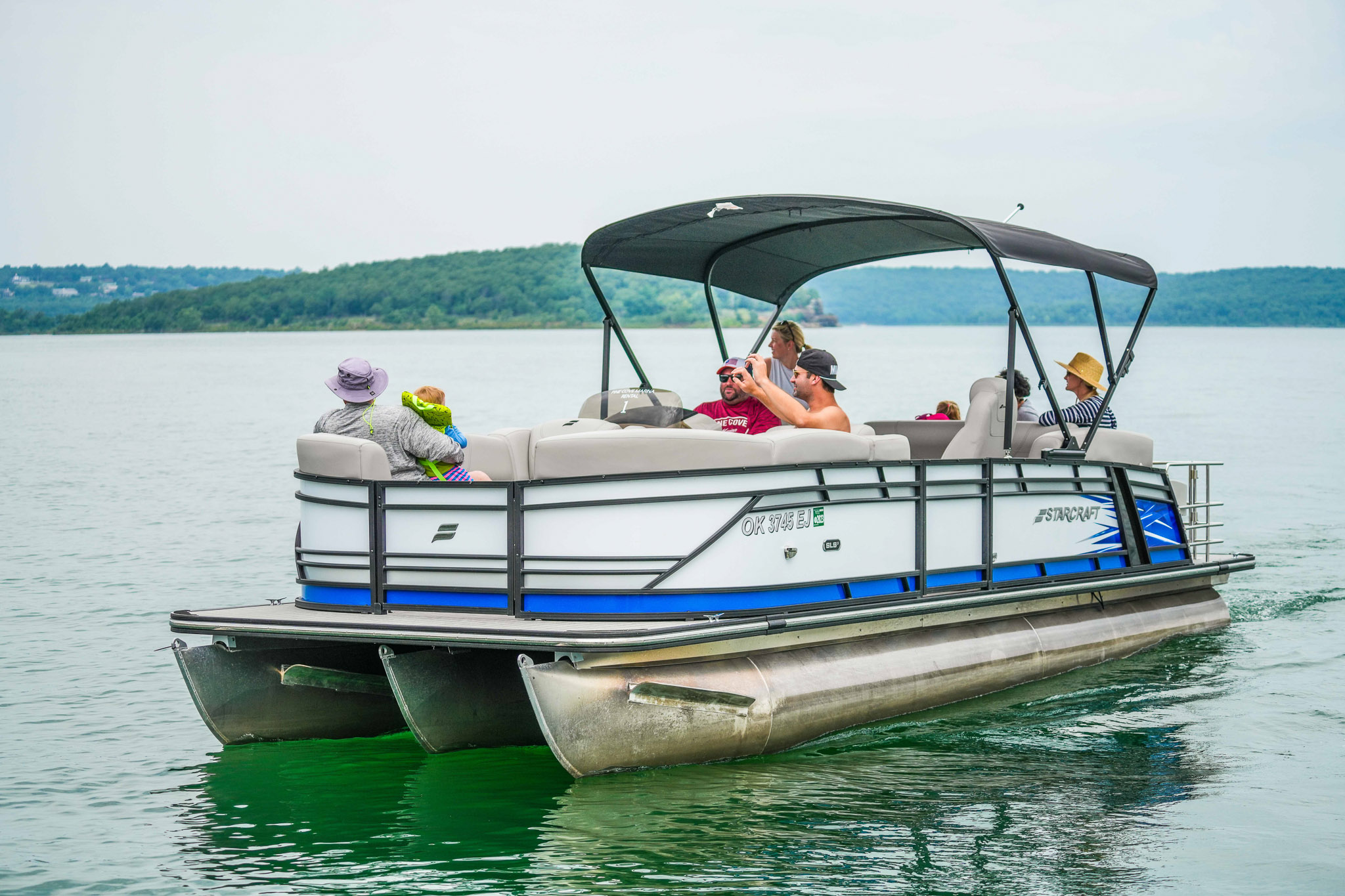 A group of people enjoy a leisurely ride on a pontoon boat at Lake Tenkiller. Seated under a canopy, theyre surrounded by calm water. Green, tree-lined hills are visible in the background, and the sky is overcast—perfect for an afternoon from Bluewater Bay Marinas boat rental service.