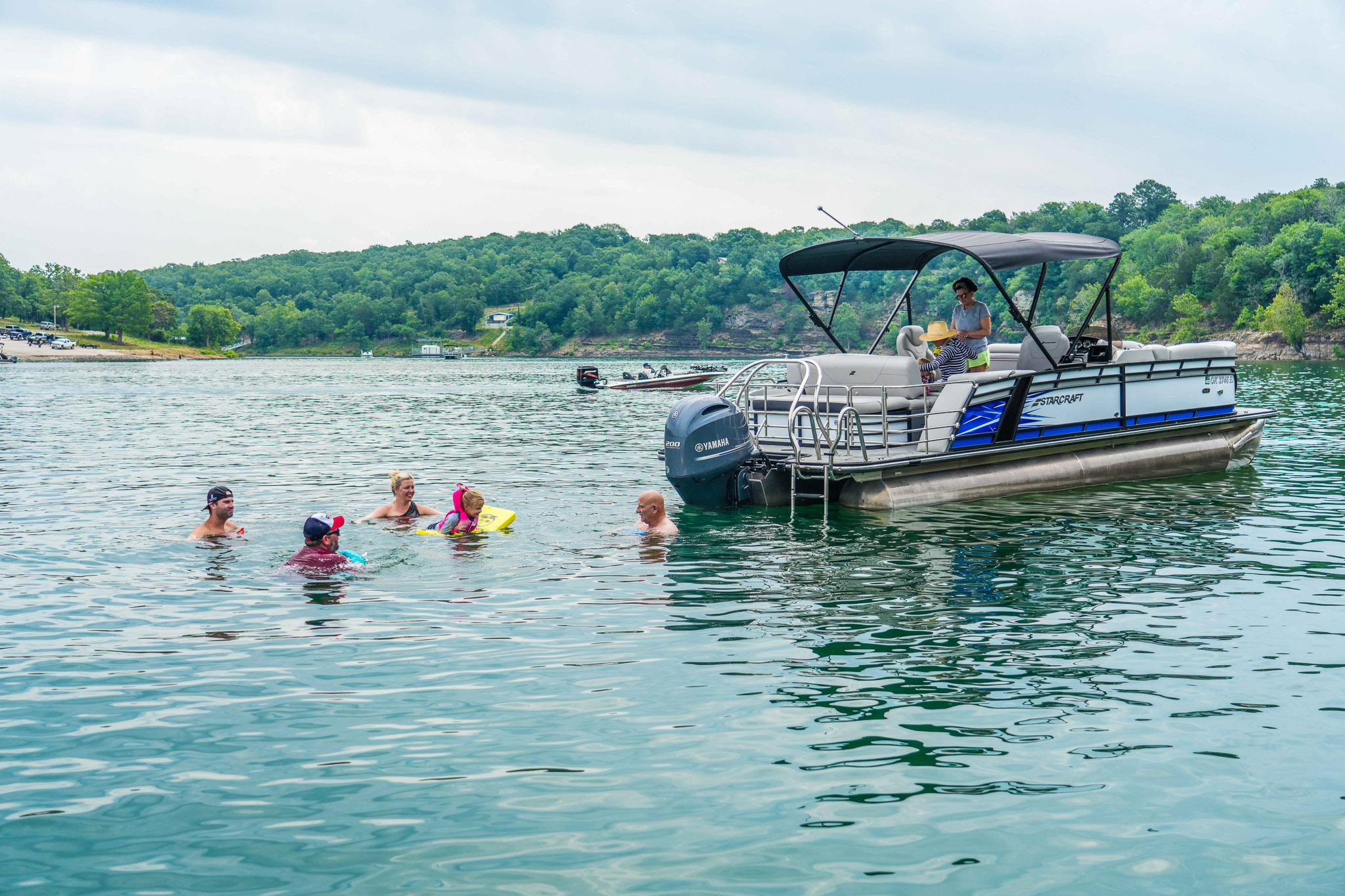 A pontoon boat, possibly from a local boat rental, is anchored on Lake Tenkiller near a wooded shore. Five people swim and float nearby, savoring a peaceful day. The sky is cloudy, and the calm water enhances the serene atmosphere at Bluewater Bay Marina.