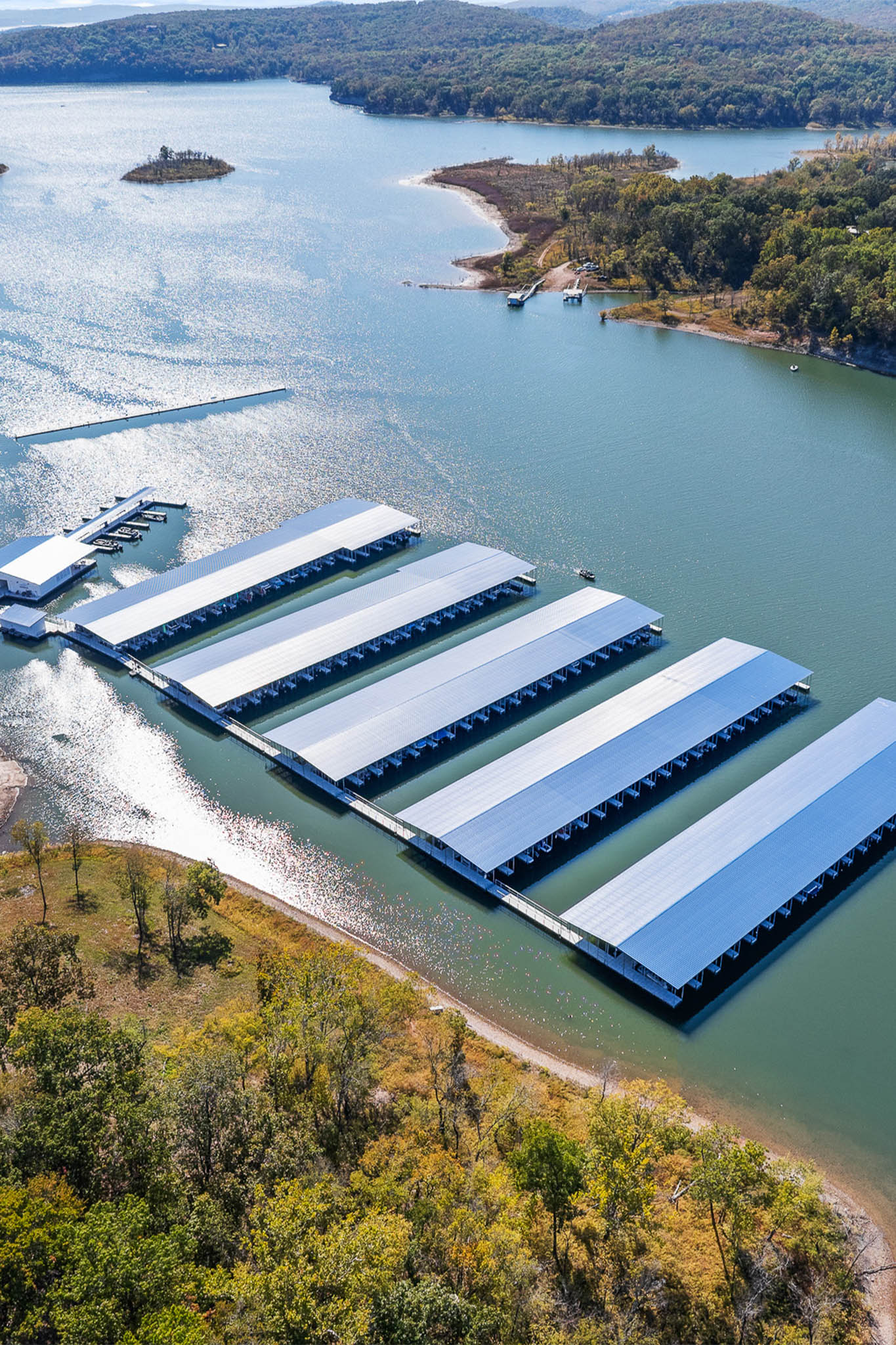 Aerial view of Lake Tenkiller with boat docks and long metal-roofed structures extending into the water at Bluewater Bay Marina. The shoreline, perfect for boat rental adventures, is surrounded by trees, with small islands visible under a clear sky.